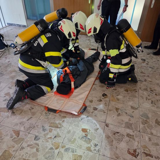 Drei Feuerwehrleute in voller Ausrüstung führen eine Rettungsaktion an einer Person durch, die auf einer Trage liegt. Die Person ist mit einem Gurt gesichert. Die Szene spielt sich drinnen ab, mit gefliesten Böden und einer Glastür im Hintergrund.
