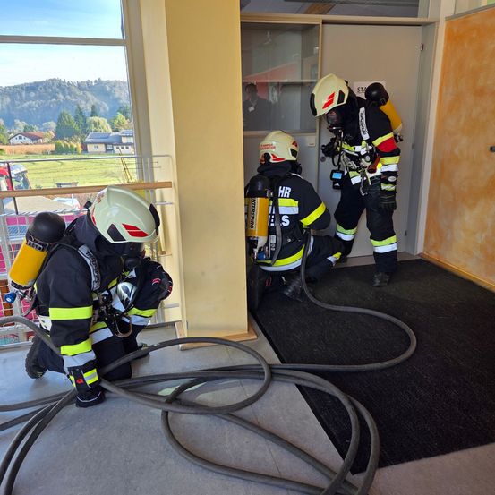 Drei Feuerwehrleute arbeiten in einer Trainingsumgebung, einer betritt eine Tür, ein anderer sitzt und hält Ausrüstung, und der dritte kniet und handhabt einen Schlauch.