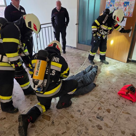 Eine Gruppe von Feuerwehrleuten in Schutzkleidung übt eine Rettungsaktion auf einem Boden, wobei eine Person liegt und eine andere ein Seil hält.