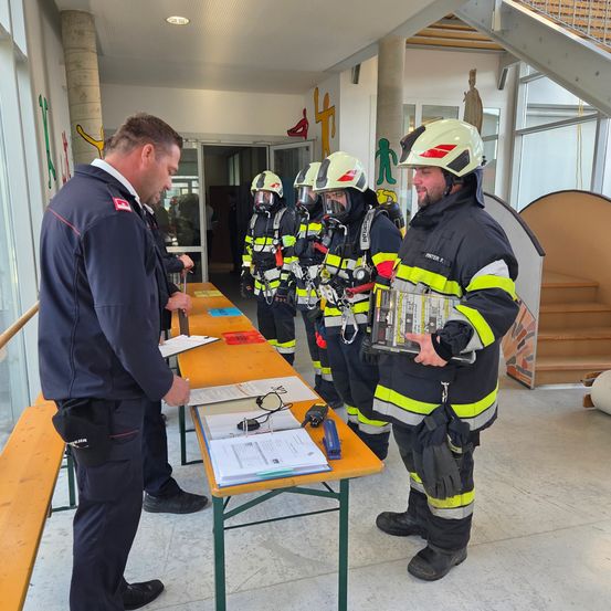 Mehrere Feuerwehrleute in voller Ausrüstung stehen in einer Reihe in einer Halle, während ein Offizier mit ihnen spricht. Papiere und ein Gerät liegen auf einem Tisch vor ihnen.