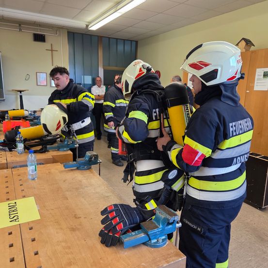 Eine Gruppe von Feuerwehrleuten arbeitet in einem Raum mit Ausrüstung. Einer justiert einen Helm, während ein anderer einen Feuerlöscher hält. Sie tragen die volle Ausrüstung.