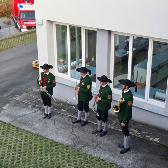 Vier Männer in grünen Uniformen und Hüten stehen in einer Reihe und spielen Trompeten vor einem Gebäude mit Glasfenstern. Ein Feuerwehrauto ist in der Nähe geparkt.