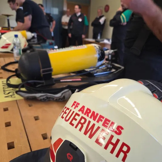 Eine Gruppe von Menschen in einem Raum mit Feuerwehrausrüstung auf einem Tisch. Helme und Flaschen sind sichtbar. Ein Helm sagt FF und FEUERWEHR.