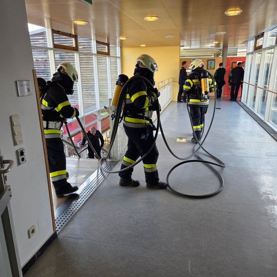 Drei Feuerwehrleute üben in einem Flur mit Schläuchen. Sie tragen Helme und Uniformen. Hinter ihnen steht eine weitere Person auf der Treppe.