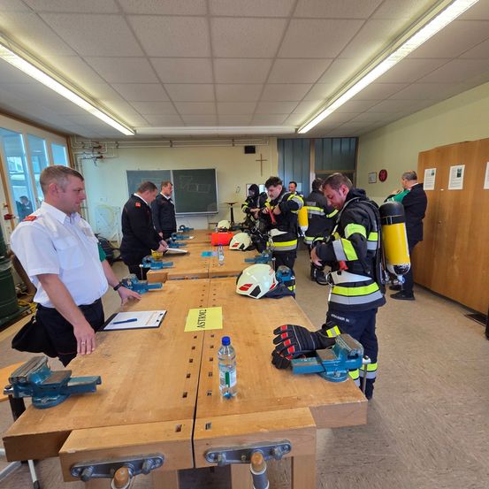 Eine Gruppe von Feuerwehrleuten ist in einem Raum versammelt, mit langen Holztischen. Helme, Werkzeuge und ein Feuerlöscher sind auf den Tischen abgestellt. Ein Mann in einem weißen Hemd steht vor einem Tisch.