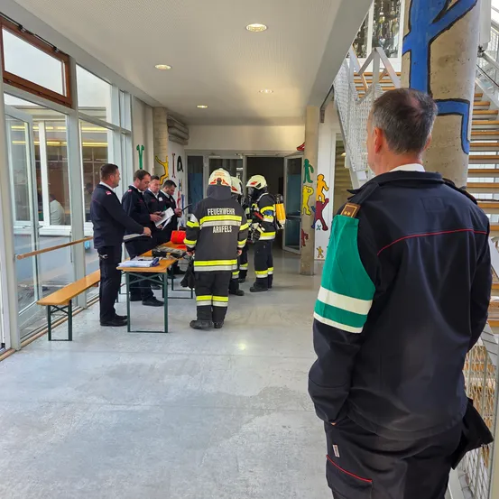 Eine Gruppe von Feuerwehrleuten in Uniformen bespricht etwas an einem Tisch in einem Korridor. Ein anderer Feuerwehrmann steht hinter ihnen.