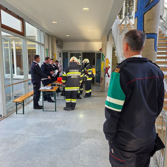 Eine Gruppe von Feuerwehrleuten in Uniformen bespricht etwas an einem Tisch in einem Korridor. Ein anderer Feuerwehrmann steht hinter ihnen.