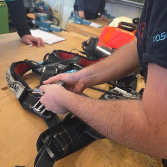 Eine Person justiert einen Sicherheitsgurt auf einem Holztisch und trägt ein Hemd mit einem Logo. Eine weitere Person ist im Hintergrund zu sehen, und verschiedene Werkzeuge und Ausrüstungsgegenstände sind vorhanden.