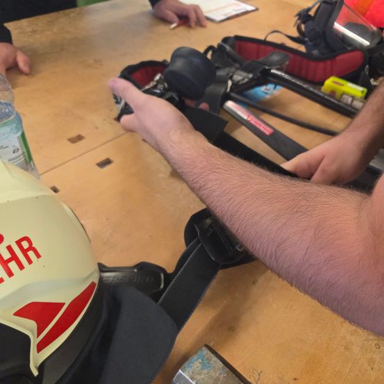 Eine Person justiert einen Sicherheitsgurt, während sie an einem Holztisch mit anderen Sicherheitsausrüstungen und einer Flasche sitzt.