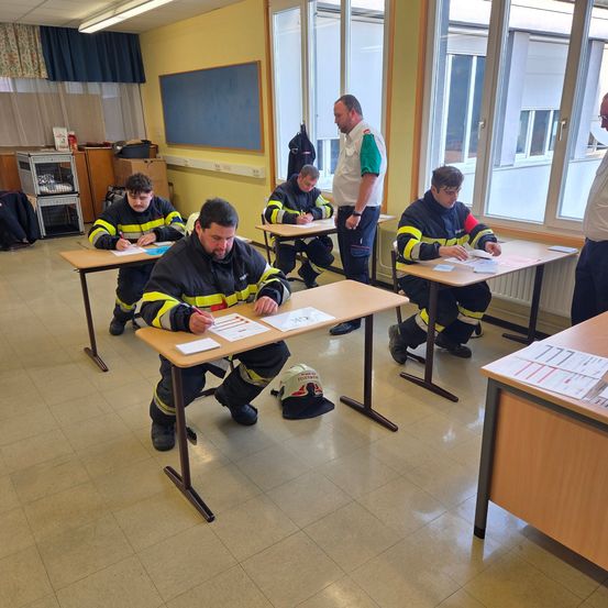 Ein Klassenzimmer mit gelben Wänden, Tischen und einer Gruppe von Feuerwehrleuten, die eine schriftliche Prüfung ablegen. Ein Mann in einem weißen und grünen Uniform steht vorne.