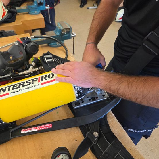 Eine Person repariert eine gelbe Maschine mit 'INTERSPIRO' darauf. Die Maschine steht auf einem Holztisch, und es gibt andere Werkzeuge in der Nähe.