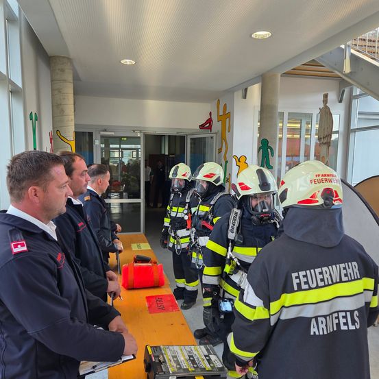Eine Gruppe von Feuerwehrleuten in voller Ausrüstung, einschließlich Helme und Jacken, steht in einem Korridor. Sie werden von einem Mann mit einem Klemmbrett eingewiesen. In der Nähe steht ein Tisch mit verschiedenen Ausrüstungsgegenständen, einschließlich einer roten Tasche und einer schwarzen Box.