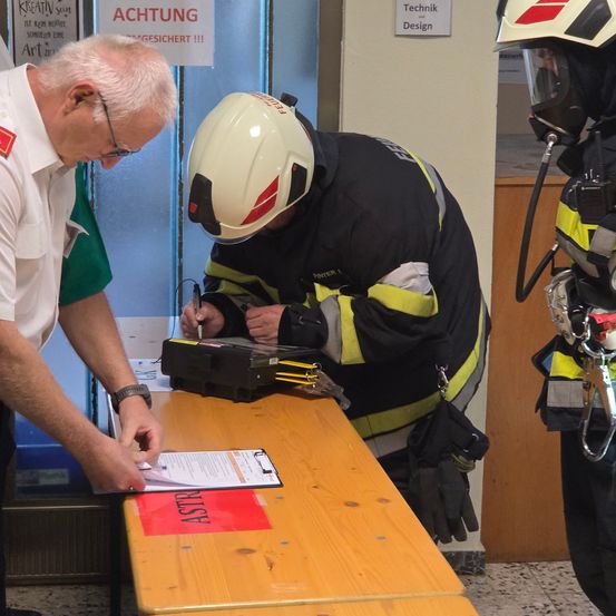 Ein älterer Mann in einem weißen Hemd unterzeichnet ein Dokument an einem Schreibtisch mit einem Feuerwehrmann in voller Ausrüstung. Ein weiterer Feuerwehrmann in voller Ausrüstung steht in der Nähe.