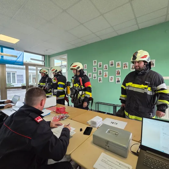 Fünf Feuerwehrleute in Helmen stehen neben einem Tisch mit Papieren und einem Laptop, während ein anderer Mann auf Papier schreibt.