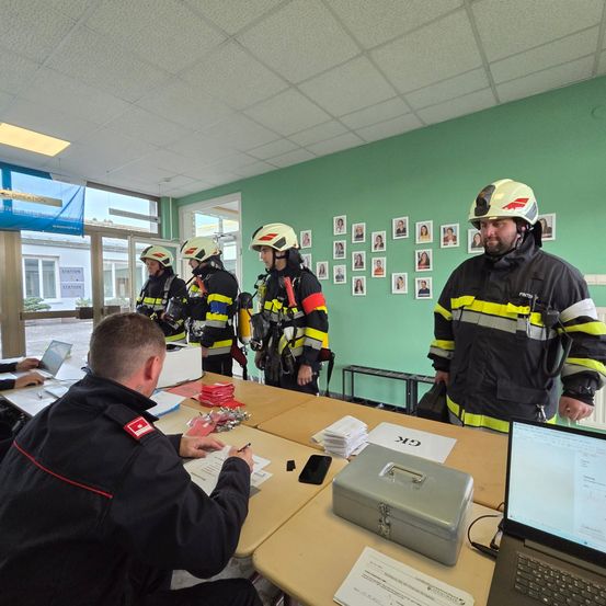 Fünf Feuerwehrleute in Helmen stehen neben einem Tisch mit Papieren und einem Laptop, während ein anderer Mann auf Papier schreibt.