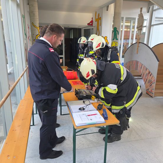 Mehrere Feuerwehrleute in Schutzkleidung arbeiten an einem Tisch, wobei eine Person auf einen Laptop konzentriert ist und eine andere hinter ihm steht. Sie befinden sich in einem Raum mit einer Glaswand, Bänken und farbenfrohen Wanddekorationen.