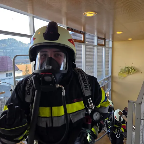 Ein Feuerwehrmann in voller Ausrüstung, einschließlich Helm und Gasmaske, steht in einem Gebäude mit großen Fenstern. Ein weiterer Feuerwehrmann steht hinter ihm, ebenfalls ausgerüstet. Die Szene wird von Deckenleuchten beleuchtet und enthält einen dekorativen Ventilator an der Wand.