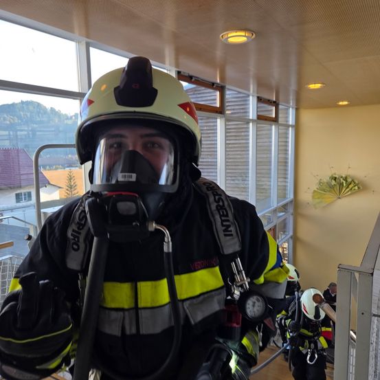 Ein Feuerwehrmann in voller Ausrüstung, einschließlich Helm und Gasmaske, steht in einem Gebäude mit großen Fenstern. Ein weiterer Feuerwehrmann steht hinter ihm, ebenfalls ausgerüstet. Die Szene wird von Deckenleuchten beleuchtet und enthält einen dekorativen Ventilator an der Wand.