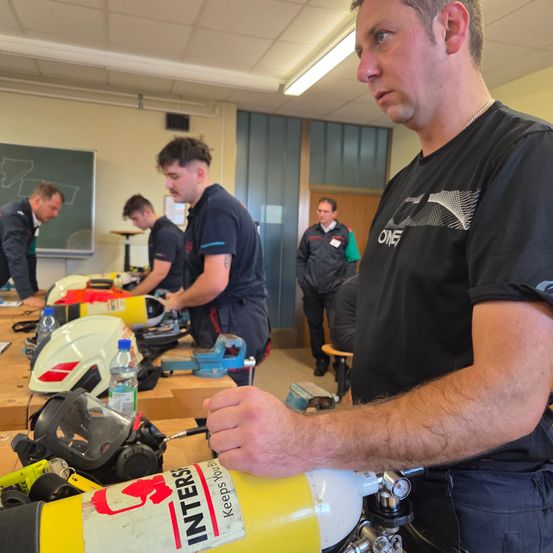 Mehrere Personen arbeiten in einer Werkstatt. Ein Mann in einem schwarzen T-Shirt hält eine gelbe Sauerstoffflasche. Andere Personen stellen Ausrüstung auf dem Tisch ein. Ein Tafel ist im Hintergrund sichtbar.