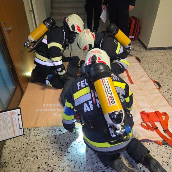 Mehrere Feuerwehrleute führen eine Rettungsaktion durch. Einer liegt auf dem Boden, während andere knien und Gasmasken tragen. Sie haben Sauerstoffflaschen auf dem Rücken.