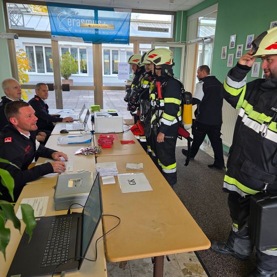 Mehrere Feuerwehrleute in voller Ausrüstung stehen in einem Raum mit einem langen Tisch, Laptops und Papieren. Ein Mann berührt seinen Helm.