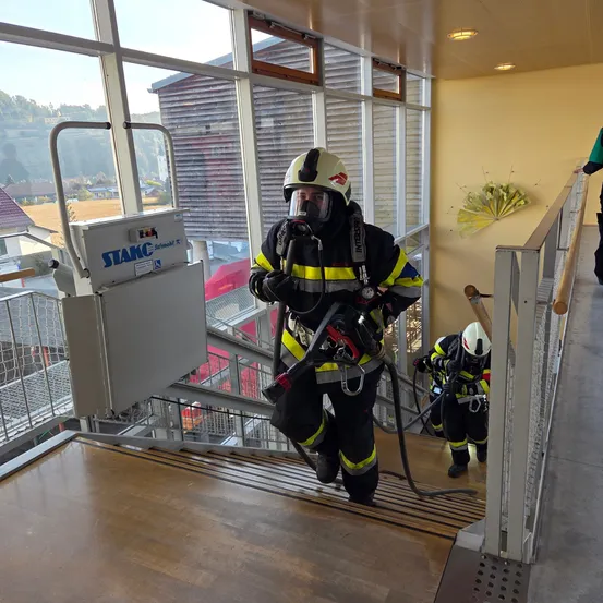 Zwei Feuerwehrleute steigen eine Treppe hinab, einer mit Maske und Schlauch, der andere folgt dicht dahinter. Die Treppe ist gut beleuchtet mit großen Fenstern und einem Metallgeländer.