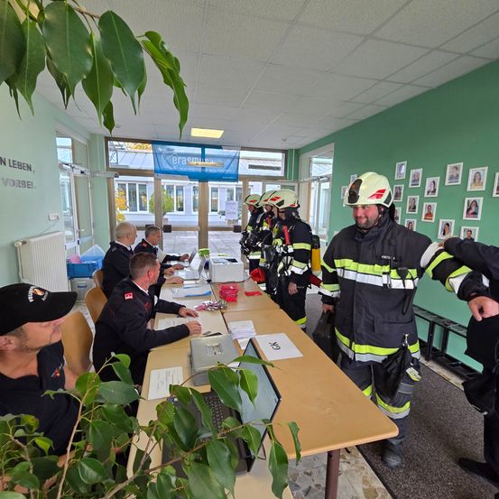 Eine Gruppe von Feuerwehrleuten ist in einem Raum mit einem Tisch versammelt. Sie tragen ihre Uniformen und Helme. Papiere und ein Laptop sind auf dem Tisch. Der Raum hat grüne Wände und Bilder an der Wand.