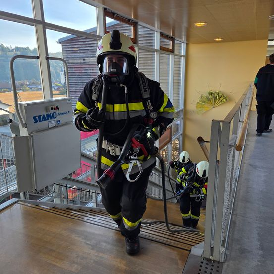 Ein Feuerwehrmann mit Atemschutzgerät geht eine Treppe hinauf und hält eine Feuerlöschanlage. Zwei weitere Feuerwehrleute folgen ihm. Die Szene spielt sich drinnen ab, mit Glasfenstern und einer gelben Wand.