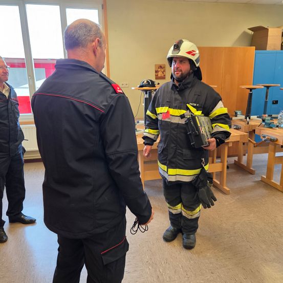 Ein Feuerwehrmann in voller Ausrüstung steht in einer Werkstatt mit einer anderen Person. Hinter ihnen sind zwei weitere Personen anwesend. Der Raum hat Holztische, einen Heizkörper und einen blauen Schrank.