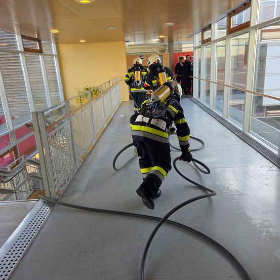 Drei Feuerwehrleute rennen mit einem Schlauch einen Flur hinunter. Einer führt den Weg. Zuschauer beobachten von der Seite.