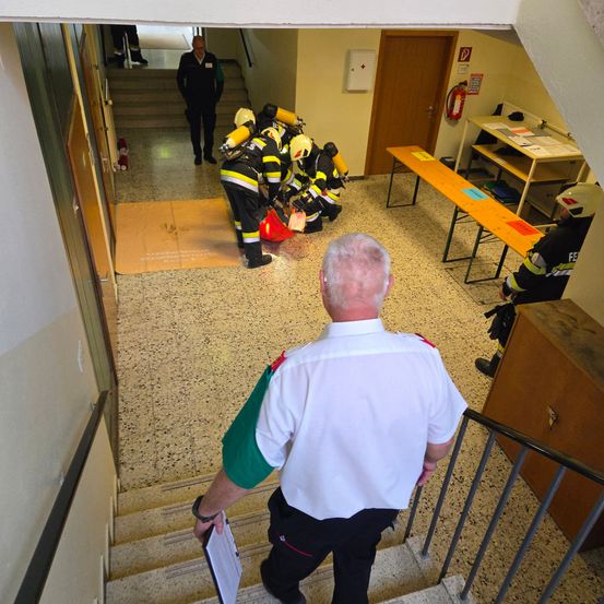Feuerwehrleute in Schutzkleidung üben Rettungsaktionen in einem Gebäudeflur. Ein Mann in weißer Uniform beaufsichtigt die Übung, während andere sitzen und stehen.