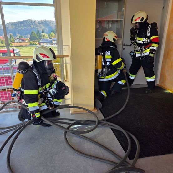 Drei Feuerwehrleute in voller Ausrüstung sind in einem Gebäude und arbeiten an einer Aufgabe. Sie benutzen Schläuche und scheinen eine Schulung durchzuführen.