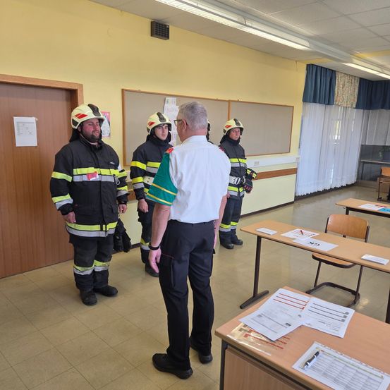 Vier Feuerwehrleute in Uniformen stehen in einem Klassenzimmer mit einem Mann in einem weißen Hemd. Papiere und Stifte sind auf den Tischen.