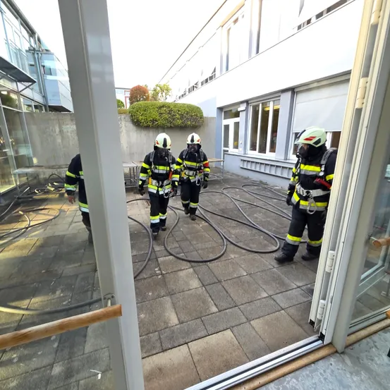 Vier Feuerwehrleute in voller Ausrüstung gehen auf einem Ziegelboden, möglicherweise für eine Trainingsübung. Ein Feuerwehrmann hält eine Feuerwehrhose. Die Szene ist im Freien in der Nähe eines Gebäudes.