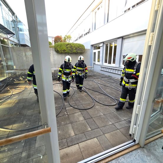 Vier Feuerwehrleute in voller Ausrüstung gehen auf einem Ziegelboden, möglicherweise für eine Trainingsübung. Ein Feuerwehrmann hält eine Feuerwehrhose. Die Szene ist im Freien in der Nähe eines Gebäudes.