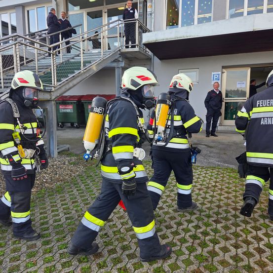 Mehrere Feuerwehrleute in voller Ausrüstung stehen vor einem Gebäude mit einer Treppe und Mülleimern. Eine Person steht in der Nähe des Eingangs.