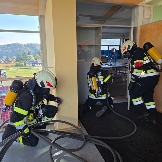 Drei Feuerwehrleute in voller Ausrüstung üben das Betreten eines Gebäudes. Einer kniet auf dem Boden, während die beiden anderen stehen, einer mit einer Schlauchleitung. Der Raum hat einen Schreibtisch und ein Fenster mit einer malerischen Aussicht.