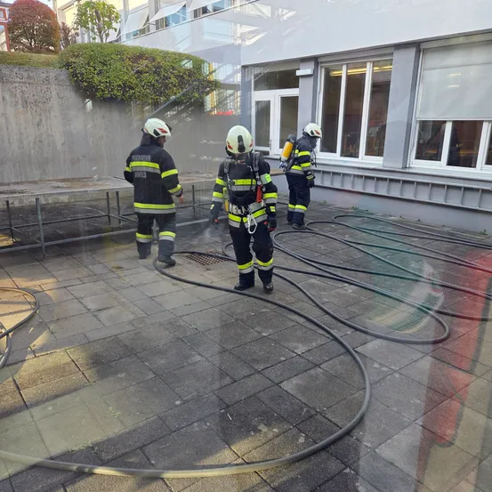 Drei Feuerwehrleute in voller Ausrüstung stehen vor einem Gebäude. Sie sprühen Wasser, wahrscheinlich als Übung für einen Brand. Große Schläuche liegen auf dem Boden.