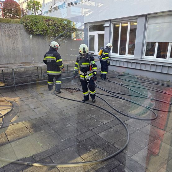 Drei Feuerwehrleute in voller Ausrüstung stehen vor einem Gebäude. Sie sprühen Wasser, wahrscheinlich als Übung für einen Brand. Große Schläuche liegen auf dem Boden.