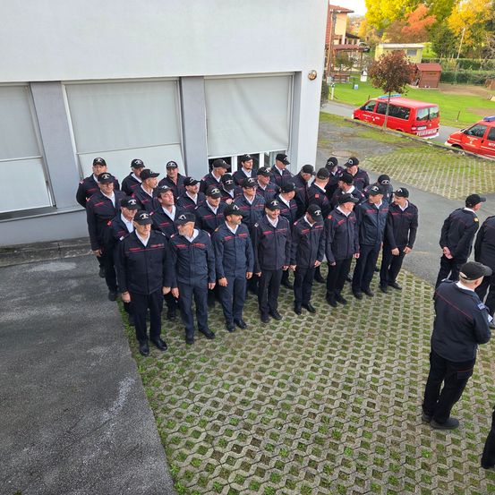 Eine Gruppe von Feuerwehrleuten in Uniform steht in einer Reihe vor einem Gebäude mit zwei roten Feuerwehrfahrzeugen, die draußen geparkt sind.