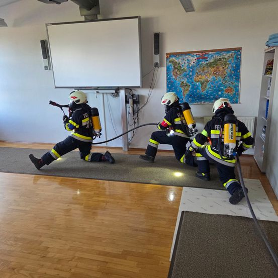 Drei Feuerwehrleute in voller Ausrüstung trainieren auf einem polierten Holzboden. Sie haben eine große Weltkarte und eine Tafel an der Wand angebracht.