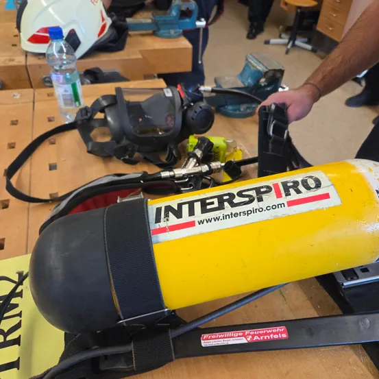 Ein gelber Sauerstofftank mit dem Label Interspiro. Ein Helm und eine Flasche sind in der Nähe. Eine Person hält den Tank in der Hand.