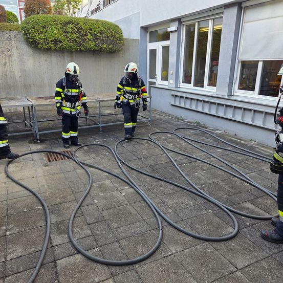 Drei Feuerwehrleute üben mit Schläuchen auf einem gefliesten Boden vor einem Gebäude mit Glasfenstern.
