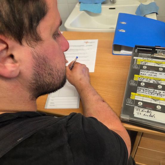 Ein Mann sitzt in einem Badezimmer an einem Schreibtisch, konzentriert auf ein Papier mit einem Stift. Ein blauer Ordner und ein Waschbecken sind hinter ihm.