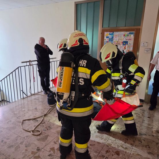 Feuerwehrleute in voller Ausrüstung bereiten sich auf einen Notfall vor. Sie stehen auf einem gefliesten Boden mit Seilen und Ausrüstung in der Nähe. Ein Mann in einem schwarzen Anzug beobachtet sie.