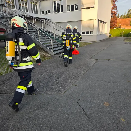 Drei Feuerwehrleute laufen auf einem Parkplatz, einer trägt einen Feuerlöscher und ein anderer eine rote Tasche. Sie sind in der Nähe eines Gebäudes mit Treppen und einem Metallzaun.