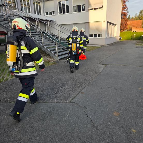 Drei Feuerwehrleute laufen auf einem Parkplatz, einer trägt einen Feuerlöscher und ein anderer eine rote Tasche. Sie sind in der Nähe eines Gebäudes mit Treppen und einem Metallzaun.