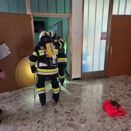 Zwei Feuerwehrleute mit Ausrüstung betreten einen Raum mit einer roten Tasche auf dem Boden. Einer hält einen Stock. Der Raum hat zwei Holztüren und eine Glastür mit Jalousien.