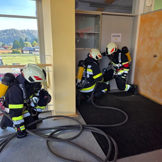 Drei Feuerwehrleute in Schutzkleidung arbeiten in einem Raum mit einer offenen Tür, die mit 'Station' beschriftet ist. Sie sind mit einem Schlauch verbunden. Durch das Fenster hat man einen Blick auf eine Landschaft mit Häusern und Bergen.