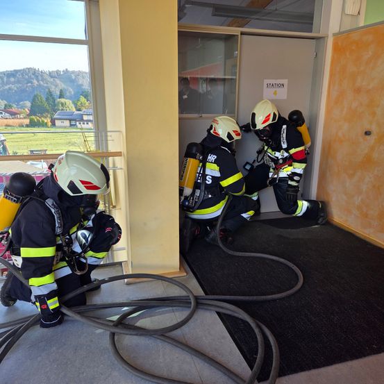 Drei Feuerwehrleute in Schutzkleidung arbeiten in einem Raum mit einer offenen Tür, die mit 'Station' beschriftet ist. Sie sind mit einem Schlauch verbunden. Durch das Fenster hat man einen Blick auf eine Landschaft mit Häusern und Bergen.